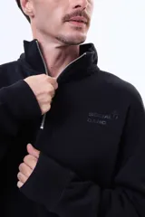 Buzo de felpa color negro con cuello alto y cierre frontal parcial. Presenta un diseño holgado con bolsillos laterales y logo bordado en el pecho.