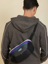 Bandolera negra con diseño técnico, confeccionada en poliamida con textura de cuadrícula. Cuenta con doble cierre frontal, bolsillos laterales de malla y correa ajustable. Incluye el logo de la marca Rusty en el frente.