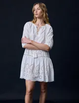 Vestido corto blanco de algodón con bordado calado tipo broderie, cuello redondo, escote en V con lazo y borlas, y mangas tres cuartos con puño.