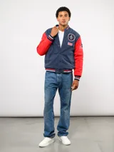 Campera bomber azul marino estilo retro, con cierre frontal de botones a presión, cuello, puños y ruedo con tejido acanalado tricolor. Presenta el logo de Umbro y el escudo del Club Nacional de Football bordados en el pecho, junto con el año 1899 y parches decorativos en las mangas.
