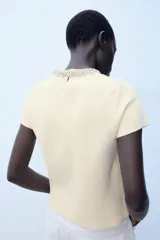Jersey de punto color amarillo claro, con cuello redondo adornado con aplicación de joyas a contraste, manga corta y cierre en la espalda con abertura y botón.