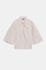 Blusa de cuello subido con escote en pico y cierre frontal con botones forrados. Presenta mangas abullonadas de largo tres cuartos con elástico en los puños y detalle de lazos en el cuello y el frente.