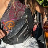 Riñonera negra con textura tipo piel de cocodrilo, con tres compartimentos con cierre y correa ajustable.