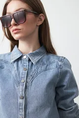 Camisa de jean color azul claro con tachas metálicas en el canesú y a lo largo de las costuras frontales. Tiene mangas largas con puños abullonados y cierre frontal con botones.