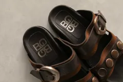 Sandalias bajas de cuero marrón con doble tira, hebilla ajustable y apliques de tachas metálicas.