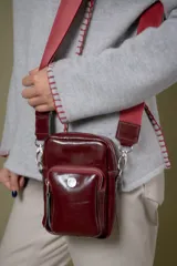 Bolso tipo pocket bag de cuero vacuno color bordó, con cierre principal y bolsillo frontal con cierre. Incluye correa de cinta regulable en color bordó y herrajes metálicos plateados.