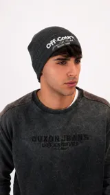 Gorro tipo beanie de punto acanalado en color gris oscuro, con bordado frontal en color blanco que incluye texto y detalles gráficos.