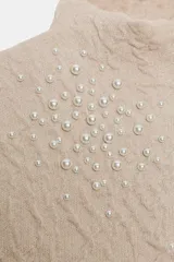 Top de punto color beige con cuello subido, manga corta y diseño texturizado con efecto arrugado. Presenta aplicaciones de perlas distribuidas en la parte frontal y las mangas.
