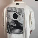 Remera negra de corte oversize con estampa rectangular en la espalda que representa una mano alcanzando un círculo sobre un fondo de líneas horizontales.