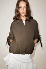 Chaqueta bomber color marrón, con cuello solapa, cierre frontal oculto con cremallera y botones a presión. Presenta mangas remangadas con trabillas y bajo abullonado con elástico interior.