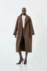 Abrigo largo tipo sobretodo de color marrón, confeccionado en una mezcla de poliéster y lana. Presenta mangas ranglán, pinzas frontales que aportan estructura y un cuello alto tipo tortuga con cierre cruzado mediante un botón.