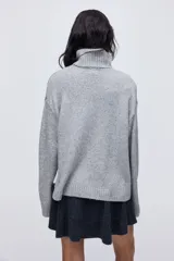 Sweater de punto gris con cuello alto acanalado, diseño jacquard frontal con paisaje de montaña y teleférico en colores blanco, negro y rojo, y hombros caídos.