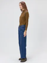 Jeans de corte ancho (wide leg) y tiro medio, confeccionados en denim de algodón negro con costuras contrastantes en color ocre. Presenta múltiples botones metálicos frontales y tachas metálicas aplicadas a lo largo de los laterales y la pretina.