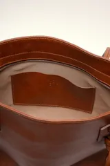 Bolso tipo tote color marrón de cuero suave, con doble asa larga decorada con nudos en los extremos inferiores y detalle de logo grabado en el frente.
