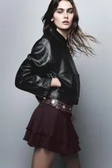 Falda pantalón de tiro medio en color marrón oscuro con acabado satinado. Presenta un diseño con volantes en el ruedo, frunces laterales y un cinturón ancho integrado con detalles de tachas metálicas doradas.