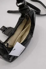 Cartera de hombro de tamaño compacto con acabado texturizado efecto cuero desgastado. Presenta un diseño con cierre superior, bolsillos frontales con cierre y una solapa central decorativa con hebilla metálica. Incluye asa fija para llevar al hombro.