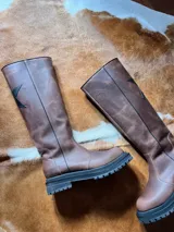 Botas de caña alta color beige y puntera marrón, con suela track y detalle de estrella blanca en el lateral.