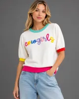 Sweater de punto de manga corta en color blanco, con cuello redondo en color turquesa, puños en colores amarillo y rojo, y borde inferior en color fucsia. Presenta la palabra 'cowgirls' bordada en relieve con letras multicolores.