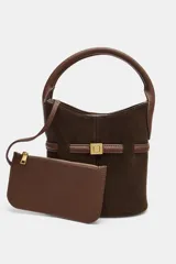 Bolso tipo saca de tamaño mini, confeccionado en gamuza color marrón oscuro con detalles en cuero marrón. Presenta un asa de mano fija, una tira decorativa horizontal con hebilla metálica dorada en el frente y costuras verticales visibles.