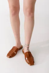 Zueco de cuero color tostado con diseño de tiras anchas entrelazadas en el empeine, punta cuadrada y suela plana.