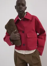 Chaqueta corta de algodón en color rojo, con cuello camisero, manga larga y cierre frontal de botones oscuros. Presenta dos bolsillos con solapa en el frente.