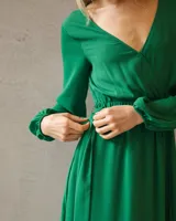 Vestido largo de satén color verde esmeralda, con escote cruzado, mangas largas amplias con puños elásticos y cintura marcada con lazo ajustable.