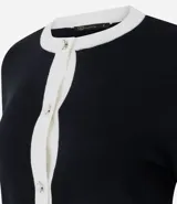 Cardigan de punto color blanco con detalles en negro en cuello, puños y tapeta frontal. Presenta cierre frontal con botones perlados y dos bolsillos delanteros falsos con solapa bicolor.