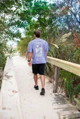 Remeron de corte oversize color violeta lavado, con estampa gráfica en la espalda que representa un paisaje natural con palmeras y un río, junto con el texto 'Part of Nature like you' y 'Glassy Waves x Origen'.