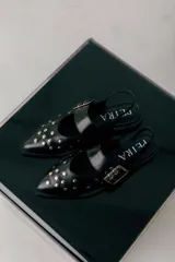 Chatas de cuero negro con punta fina, decoradas con tachas metálicas plateadas. Presentan una tira con hebilla ajustable sobre el empeine y diseño destalonado con tira trasera.