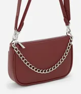 Cartera tipo baguette color bordó, confeccionada en poliuretano con acabado liso. Presenta una cadena decorativa plateada en el frente, cierre superior con cremallera y correa larga ajustable y desmontable.