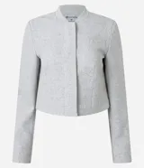 Chaqueta de sastre estructurada color gris claro, con cuello redondo tipo rectilínea, cierre frontal con botones y mangas largas con botones en los puños. Presenta un corte levemente corto y diseño minimalista.