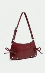 Cartera tipo baguette de charol color bordó, con diseño estructurado, cierre superior y detalles de tachas metálicas en las esquinas inferiores. Incluye tiras laterales decorativas y correa de hombro ajustable.