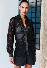 Camisa negra de encaje con detalles en cuero sintético, que incluyen el cuello, la tapeta de botones a presión, los bolsillos frontales con solapa, los puños y el borde inferior. Presenta mangas largas y un diseño estructurado y sofisticado.