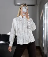 Camisa blanca de estilo romántico con cuello alto, cierre frontal de botones y detalles de encaje y alforzas verticales. Presenta mangas largas con puños decorados con encaje.