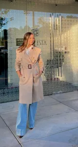 Trench coat color beige de corte clásico, con solapas amplias, doble botonadura frontal, cinturón ajustable con hebilla y trabillas en los puños.