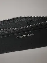 Tarjetero Calvin Klein color gris oscuro con textura de piel de cocodrilo sintética grabada. Cuenta con cuatro ranuras para tarjetas, dos bolsillos con ranura y un bolsillo monedero con fuelle y cremallera. El logo de Calvin Klein es de metal plateado brillante en la parte delantera.