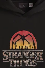 Canguro negro con capucha y bolsillo frontal tipo canguro. Presenta un estampado gráfico en el pecho con el logo de la serie Stranger Things y una silueta del Mind Flayer sobre un atardecer.