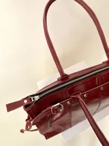 Bolso grande de cuero marrón con textura tipo cocodrilo, estructura rígida y asas para llevar al hombro. Tiene una tira con tachas y hebillas en cada extremo, también lleva lazos en los costados.