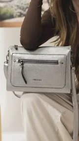 Bandolera de tamaño mediano en color gris, confeccionada en símil cuero con textura granulada. Presenta un bolsillo frontal con cierre, herrajes metálicos oscuros y correa ajustable y desmontable.