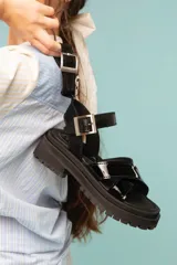 Sandalias negras de estilo chunky con tiras cruzadas en charol y base de goma dentada. Cuentan con una tira superior desmontable y cierre con hebilla metálica en el tobillo.