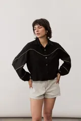 Camisa de manga larga color negro, con cuello camisero, cierre frontal con botones y detalles de ribetes contrastantes en color dorado. Presenta paneles superiores con textura y mangas abullonadas.