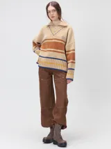 Pullover de punto con textura aterciopelada, diseño oversize, cuello polo con cierre frontal parcial y detalles de pespuntes en contraste. Presenta rayas horizontales en tonos tierra y un parche bordado con la letra M en el pecho.