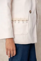 Blazer cruzado color beige, con cierre de cuatro botones frontales. Presenta detalles artesanales de encaje con perlas y cristales en la solapa y en los bolsillos delanteros.