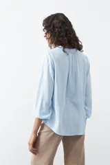 Camisa de color celeste claro con silueta relajada y mangas largas. Presenta un cuello redondo con pequeña abertura frontal y botones, además de un delicado trabajo de tablas en los hombros que aporta textura al diseño.