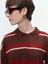 Buzo tejido de punto a rayas horizontales en tonos marrón, rojo y crema, con cuello polo y cierre frontal de botones. Presenta mangas largas con puños elastizados y un corte holgado.
