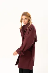 Trench de corte holgado color bordó, con cuello camisero, cierre frontal con botones y cinturón ajustable con hebilla. Presenta mangas largas con detalles de correas en los puños y bolsillos laterales.