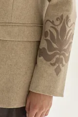 Blazer de paño color beige, con solapas clásicas y bordados decorativos en tono más oscuro en la parte inferior de las mangas.
