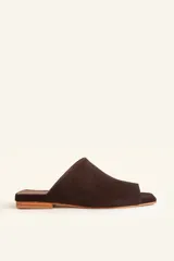 Sandalia mule de gamuza marrón, de una sola pieza con corte asimétrico y punta cuadrada, sin tiras ni hebillas, con suela plana de cuero natural.