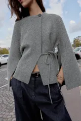 Chaqueta de punto gris con cuello redondo, manga larga acampanada y cierre frontal cruzado con botón y lazada lateral.