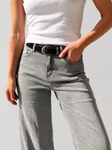 Pantalón de jean estilo palazzo, color gris claro, con corte amplio y fluido. Presenta un dobladillo ancho fijo en la botamanga, cierre frontal con botón y cremallera, y trabillas para cinturón. Incluye un cinturón negro con hebilla metálica.